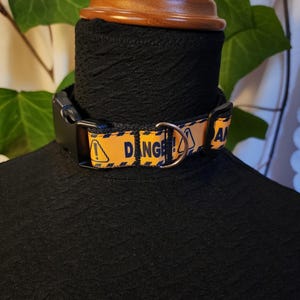 May include: A black dog collar with a yellow and black danger pattern. The collar has a black plastic buckle and a metal D-ring. The word "DANGER" is printed on the collar.