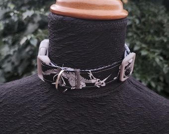 Puede incluir: Collar negro para perros con un diseño de esqueleto de dinosaurio blanco. El collar tiene una hebilla de plástico gris y un anillo de metal plateado. El collar se muestra en una cabeza de maniquí negra.