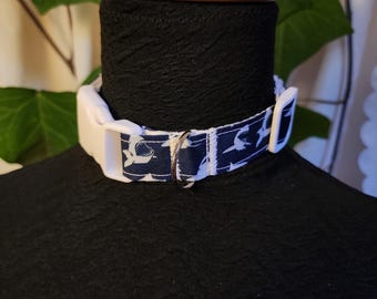 May include: A navy blue and white patterned dog collar with a white plastic buckle and a silver metal ring. The collar is displayed on a black form with a wooden top.
