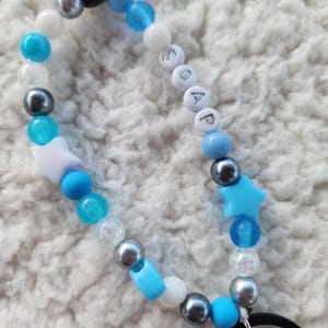 May include: A beaded keychain with a mix of blue, white, and silver beads. The word "SOAP" is spelled out with white letter beads. Features include star-shaped and round beads, and a black ring.