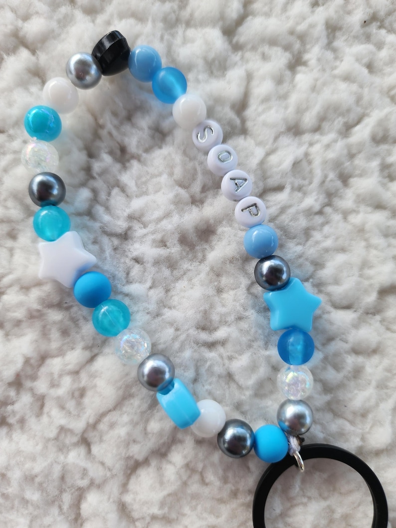 May include: A beaded keychain with a mix of blue, white, and silver beads. The word "SOAP" is spelled out with white letter beads. Features include star-shaped and round beads, and a black ring.