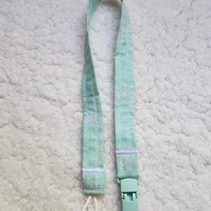 Op de afbeelding: Een lichtgroene stoffen speenclip met een witte lus en een plastic clip. De stof heeft een wit bloemenpatroon.