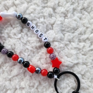 May include: A beaded lanyard with the word "GHOST" spelled out in white letter beads. The lanyard features red, black, and gray beads, a red star, and a black keyring.