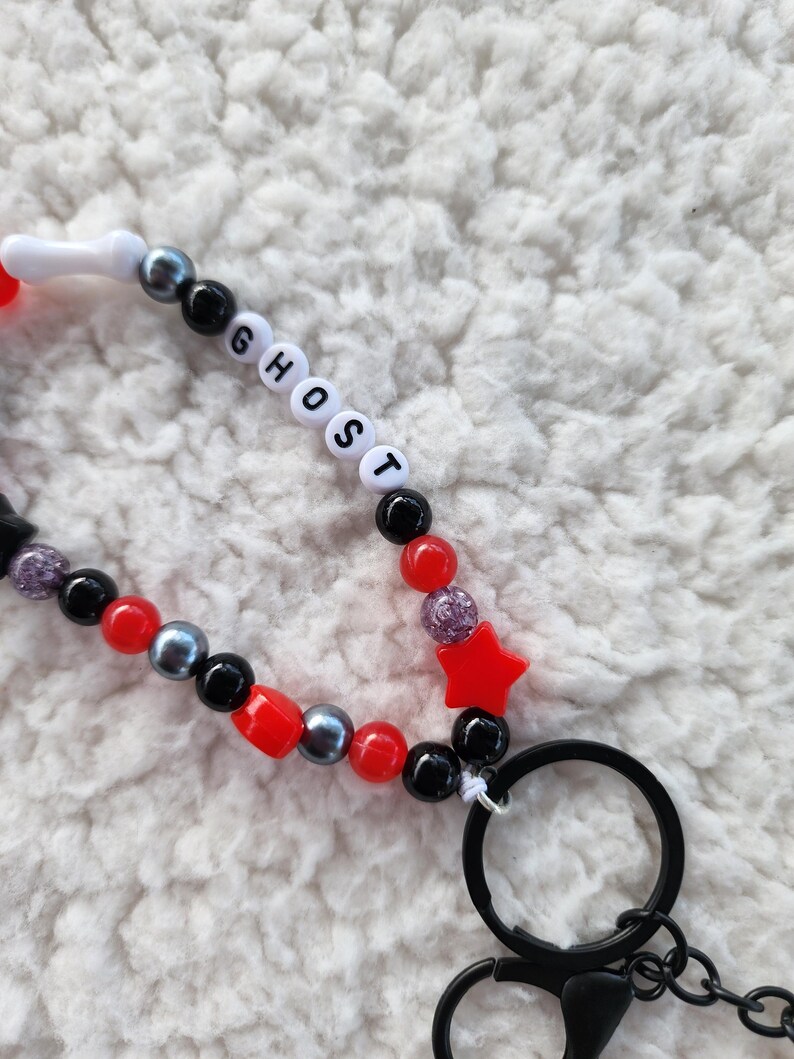 May include: A beaded lanyard with the word "GHOST" spelled out in white letter beads. The lanyard features red, black, and gray beads, a red star, and a black keyring.