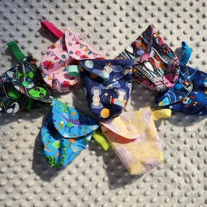 May include: A collection of seven small, fabric pouches with various colorful patterns. Designs include aliens, dinosaurs, rockets, and space themes. Each pouch has a button closure and a small loop handle, perfect for small items.