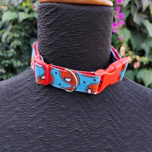 Puede incluir: Un collar de perro rojo y azul con un diseño de Spiderman. El collar tiene una hebilla de plástico roja y un anillo en D de metal plateado.