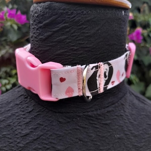 May include: Pink and white dog collar with a black and white heart pattern and pink hearts. The collar has a pink plastic buckle and a silver D-ring.
