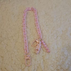Op de afbeelding: Een roze plastic ketting met een roze bedel met de tekst "SWEET" en een roze hartvormige bedel. De ketting ligt op een wit, pluche oppervlak.