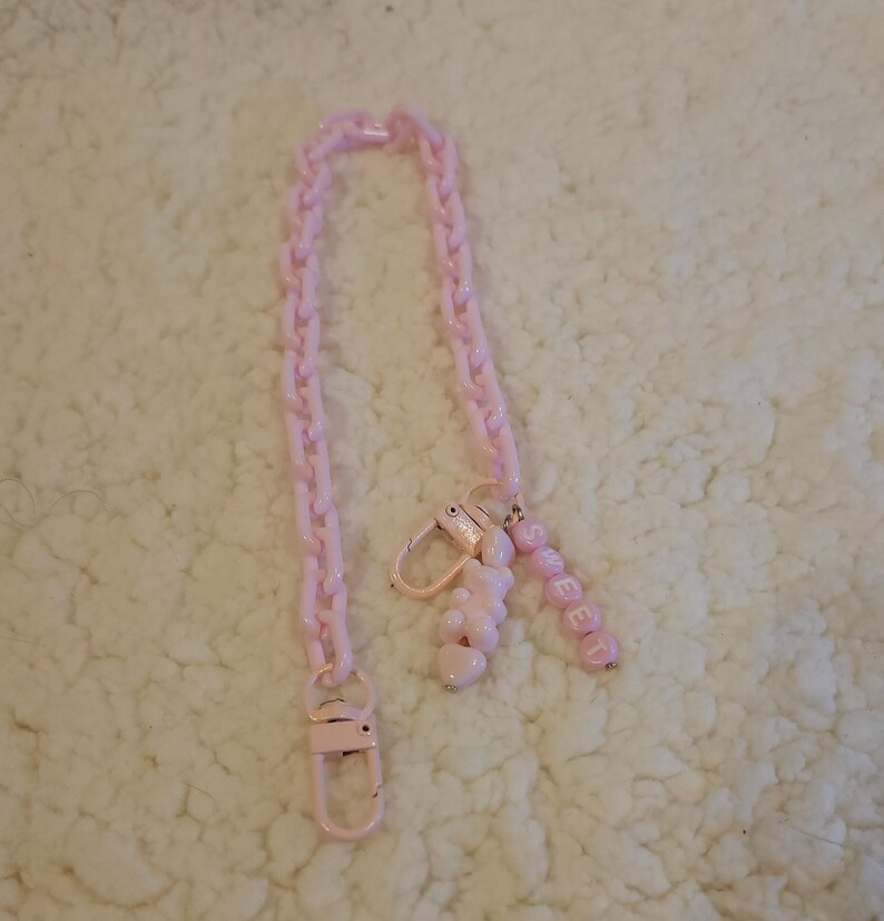 Op de afbeelding: Een roze plastic ketting met een roze bedel met de tekst "SWEET" en een roze hartvormige bedel. De ketting ligt op een wit, pluche oppervlak.