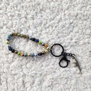 May include: A beaded keychain with the word "PRICE" spelled out in colorful beads. The keychain includes a black clasp, a metal ring, and a small gun charm. The beads are various colors, including gold, blue, and black.