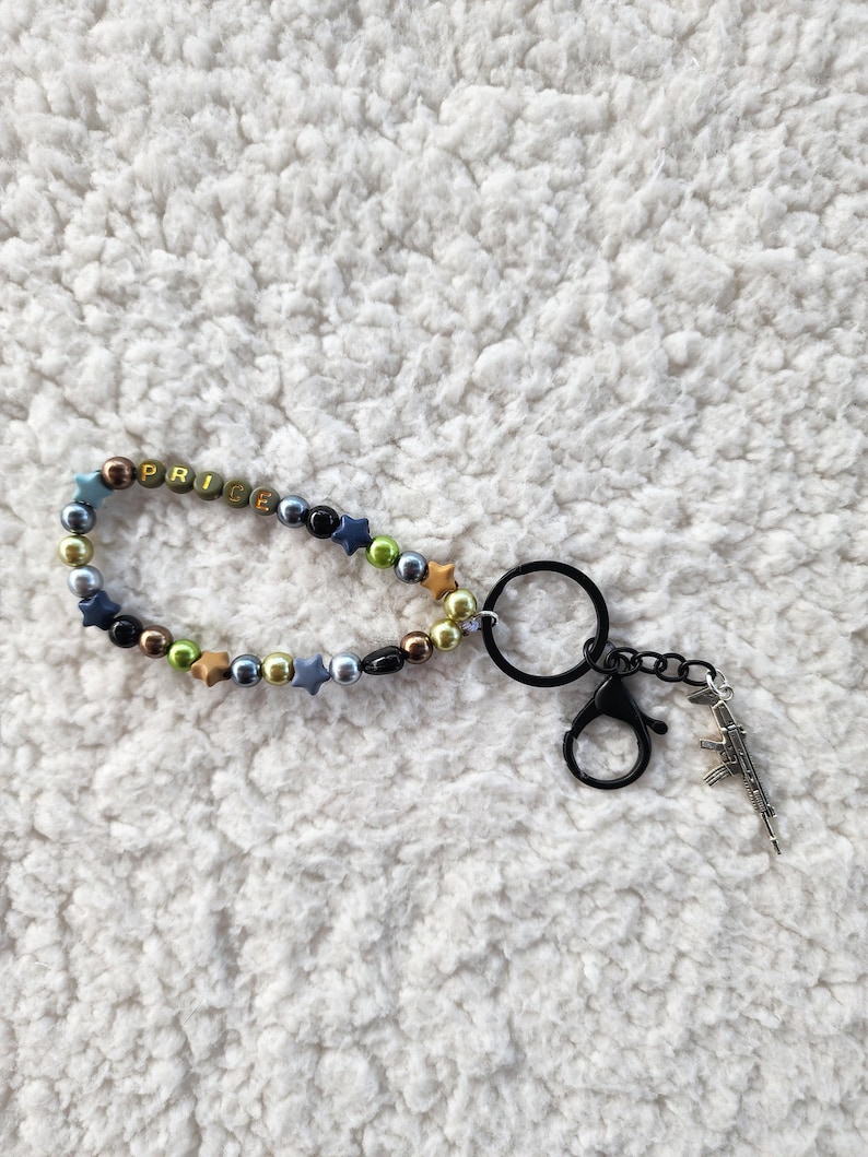 May include: A beaded keychain with the word "PRICE" spelled out in colorful beads. The keychain includes a black clasp, a metal ring, and a small gun charm. The beads are various colors, including gold, blue, and black.