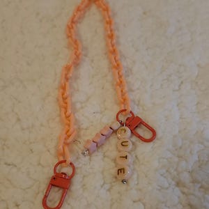 Op de afbeelding: Een roze plastic ketting met een bedel die "CUTE" zegt in gouden letters. De bedel is gemaakt van roze en witte kralen.