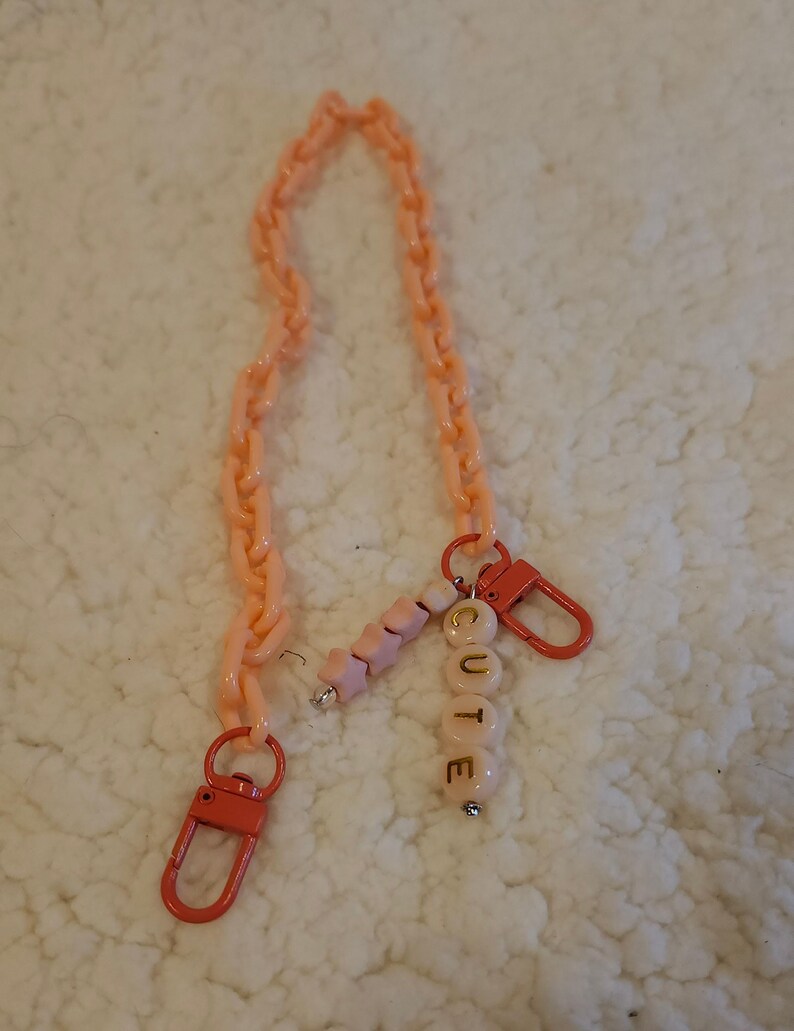 Op de afbeelding: Een roze plastic ketting met een bedel die "CUTE" zegt in gouden letters. De bedel is gemaakt van roze en witte kralen.