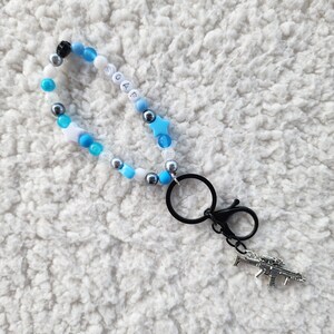 May include: A beaded keychain with the word "SOAP" spelled out in white letter beads. The keychain features blue, white, and silver beads, star-shaped beads, and a silver gun charm. It has a black clasp.