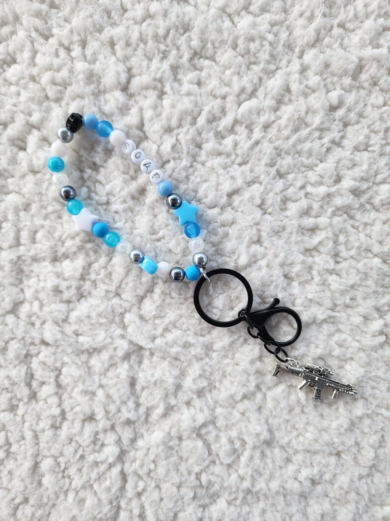 May include: A beaded keychain with the word "SOAP" spelled out in white letter beads. The keychain features blue, white, and silver beads, star-shaped beads, and a silver gun charm. It has a black clasp.