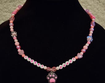 Puede incluir: Un collar de cuentas rosas con las palabras "CHEEKY PUPPY" escritas con cuentas blancas. Un dije de huella de pata cuelga del centro. El collar presenta varios tonos de cuentas rosas y detalles en forma de corazón.