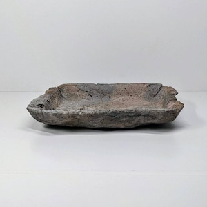 May include: A rectangular stone bowl with a rough, textured surface. The bowl has a natural colour palette of grey, brown, and pink hues. The edges are uneven, giving it a rustic appearance. Suitable for decorative purposes. Measures approximately 25 cm in length.