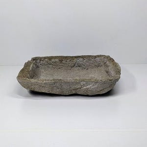 May include: A rectangular, grey stone bowl with a rough, textured surface. The bowl has a shallow interior and is likely handmade. The stone has a natural, earthy appearance, suitable for decorative or functional use.