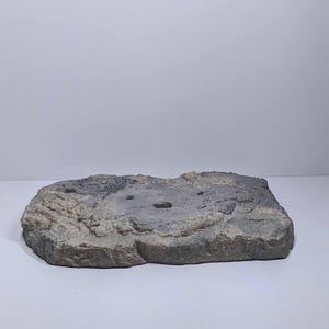 May include: A rectangular, gray, faux-stone display base with a textured surface. The base has a rough, uneven edge and three small, circular holes. The color is a mix of gray and beige.