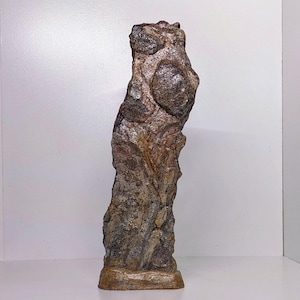 May include: A tall, textured sculpture with a rough, stone-like appearance. The sculpture features a mix of brown, gray, and tan colors, with rounded protrusions and a weathered look. It stands on a rectangular base, suggesting a decorative or artistic piece.