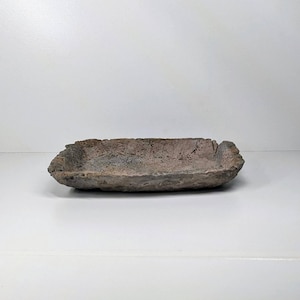 May include: A rectangular, handmade, rustic tray. The tray is a muted brown colour with a rough, textured surface. The edges have an uneven, organic shape. The tray is likely made of clay or a similar material.