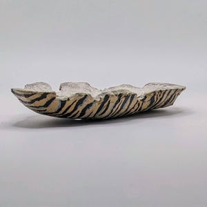 May include: A shallow, elongated ceramic dish with an irregular, hand-formed rim. The exterior features a repeating pattern of black tiger-like stripes over a light tan base, while the interior has a textured, off-white matte finish.