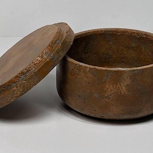 May include: A brown, round, lidded container with a textured surface. The lid is slightly ajar, revealing the interior of the vessel. The container and lid have a rustic, earthy appearance, suggesting a handmade or natural aesthetic.