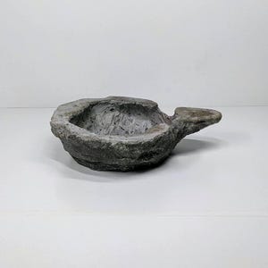 May include: A grey, textured, stone-like bowl with a handle. The bowl has a rough, uneven surface and a rounded shape. The handle extends from the side of the bowl. The bowl is likely for decorative purposes.