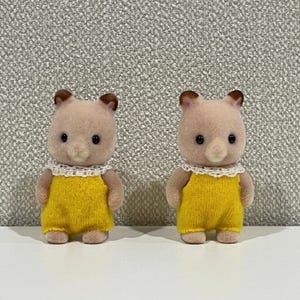 May include: Two miniature bear figurines with light brown fur, dark brown ears, and black eyes. They wear yellow overalls and white lace collars. The toys are designed for children.