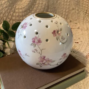 May include: A white ceramic sphere vase with a gold rim and numerous small holes. The vase is decorated with pink floral branches. The vase is sitting on top of a stack of books.