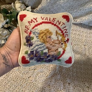 May include: A small, square, off-white needlepoint pillow with a Valentine's Day theme. The pillow features a central design of a cherub with a bow and arrow, surrounded by a red circle with the words "Be My Valentine". Accents include red hearts and purple flowers.