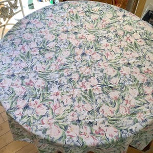 May include: A round tablecloth with a floral pattern. The design features pink, white, and blue flowers with green leaves on a white background. The tablecloth is spread over a round table.