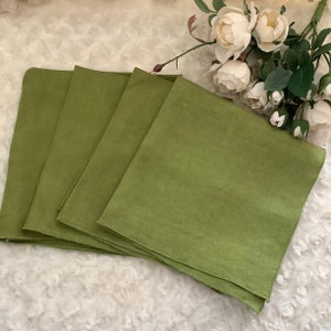 May include: Four square, olive green linen napkins are stacked on a white, textured surface. The napkins are arranged in a fan shape, with the top one slightly overlapping the others. White flowers with green stems are in the background.