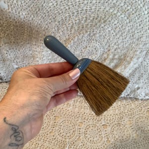 May include: A small, handheld brush with a gray handle and brown bristles. The brush is designed for cleaning and dusting. The handle is rounded and the bristles are tightly packed. The brush is held in a hand.