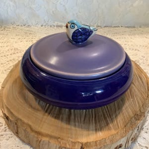 May include: A round ceramic lidded dish with a deep cobalt blue base and a lighter lavender purple lid. The lid features a small, sculpted ceramic bird figurine as a handle, painted with blue and white details and a black eye.