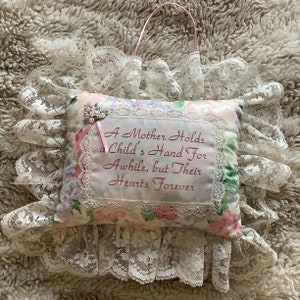 May include: A decorative pillow with a floral pattern and lace trim. The pillow has the embroidered text: "A Mother Holds a Child's Hand For Awhile, but Their Hearts Forever." A pink ribbon and a pink handle are attached.