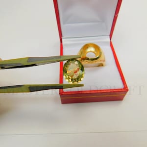 Puede incluir: Una piedra preciosa verde sostenida con unas pinzas, con un engaste de anillo de oro en el fondo.