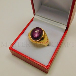 Op de afbeelding: Een gouden ring met een grote, ovale, donkerpaarse edelsteen gezet in een bezel setting. De ring bevindt zich in een rode fluwelen ringdoos.