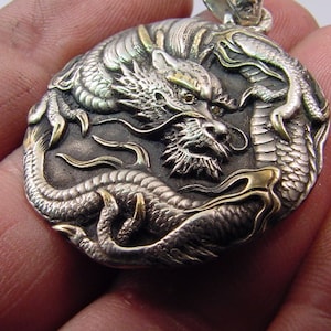 May include: Silver and gold dragon pendant with intricate details. The dragon is curled around itself with its head facing forward. The pendant is a circular shape and has a textured surface.