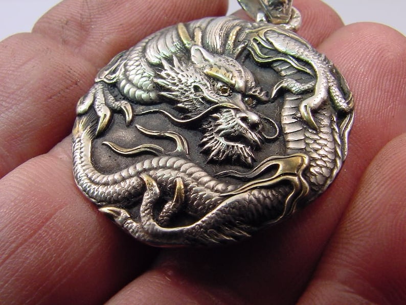 May include: Silver and gold dragon pendant with intricate details. The dragon is curled around itself with its head facing forward. The pendant is a circular shape and has a textured surface.