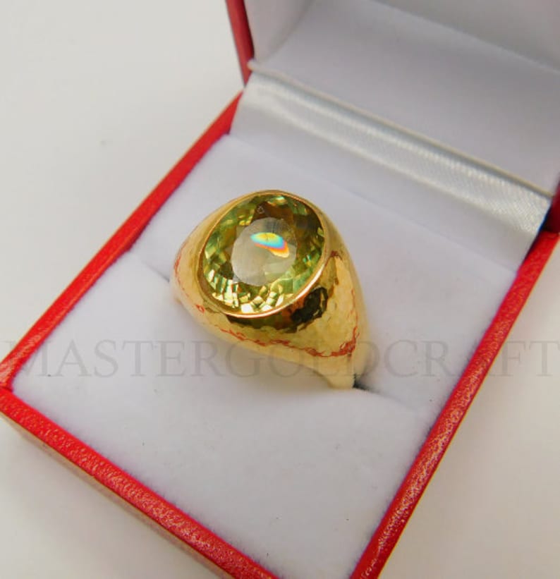 Puede incluir: Un anillo de oro con una gran piedra preciosa verde de forma ovalada. El anillo est&aacute; engastado en una banda de oro martillada.