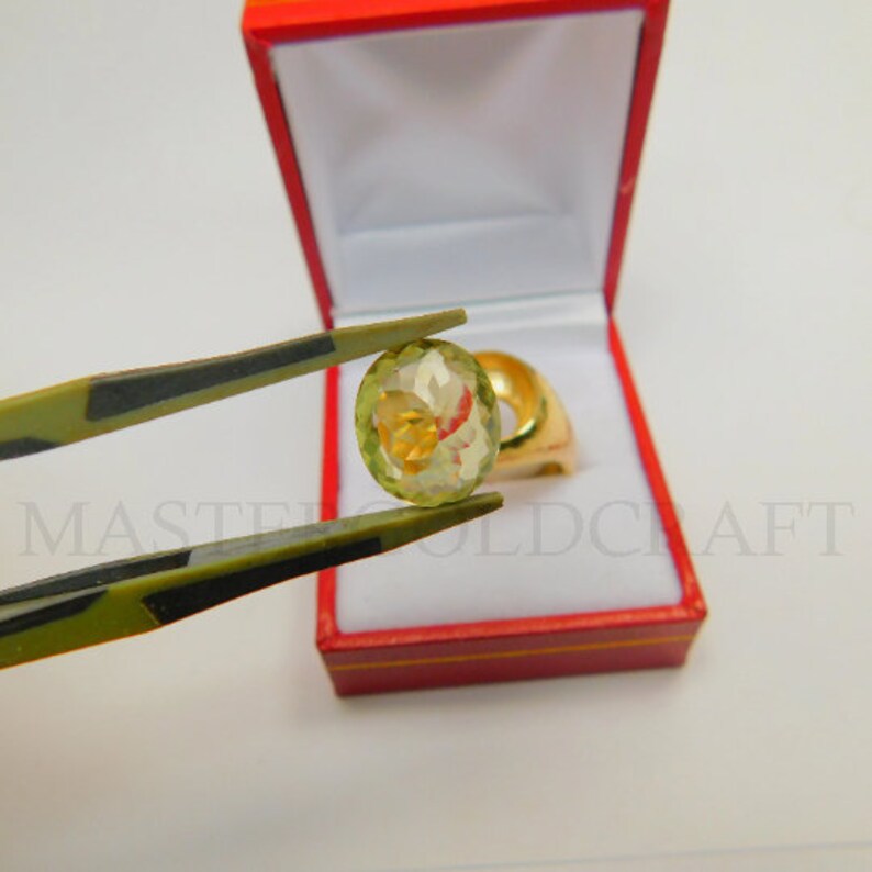 Puede incluir: Primer plano de una gema ovalada verde claro sostenida por unas pinzas. La gema est&aacute; engastada en un anillo de oro, que se encuentra en una caja de anillo de terciopelo rojo.