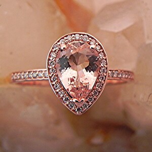 May include: A pear-shaped morganite engagement ring set in rose gold with a halo of smaller diamonds. The ring has a delicate band with small diamonds.