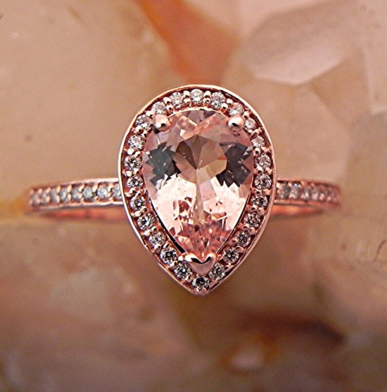 May include: A pear-shaped morganite engagement ring set in rose gold with a halo of smaller diamonds. The ring has a delicate band with small diamonds.