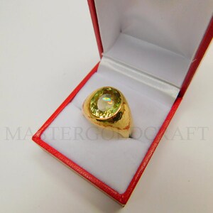 Puede incluir: Un anillo de oro con una gran piedra preciosa redonda de color verde engastada en una banda texturizada. El anillo se muestra en una caja de anillo de terciopelo rojo.
