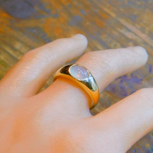 K&ouml;nnte beinhalten: Ein goldener Ring mit einem wei&szlig;en ovalen Edelstein, der in einer glatten, abgerundeten Fassung sitzt. Der Ring wird an einer Hand getragen.