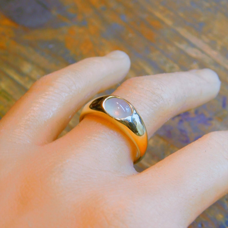 K&ouml;nnte beinhalten: Ein goldener Ring mit einem wei&szlig;en ovalen Edelstein, der in einer glatten, abgerundeten Fassung sitzt. Der Ring wird an einer Hand getragen.