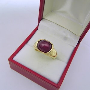 May include: A gold ring with a large, oval-shaped ruby gemstone set in a bezel setting. The ring is in a red velvet ring box.