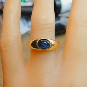 K&ouml;nnte beinhalten: Ein goldener Ring mit einem blauen Cabochon-Edelstein, der in einer Zargenfassung gefasst ist.