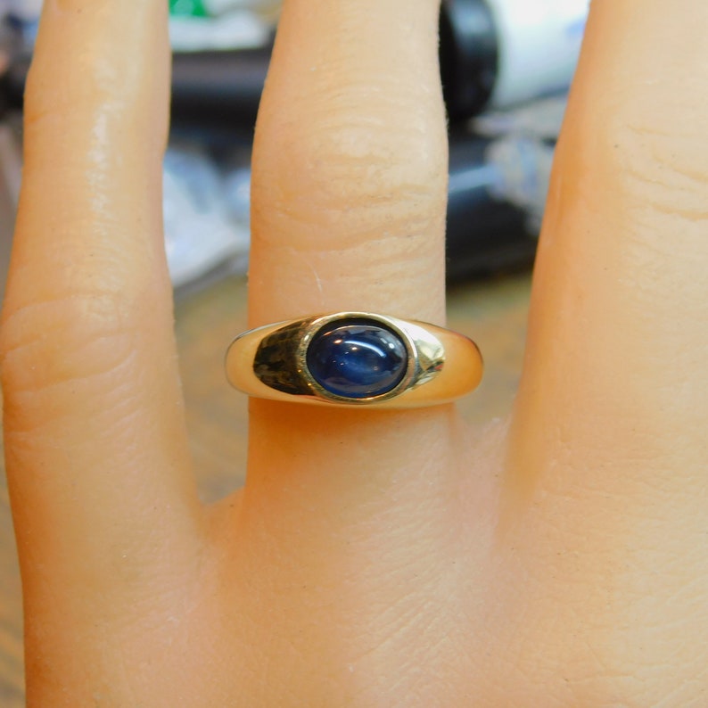 K&ouml;nnte beinhalten: Ein goldener Ring mit einem blauen Cabochon-Edelstein, der in einer Zargenfassung gefasst ist.
